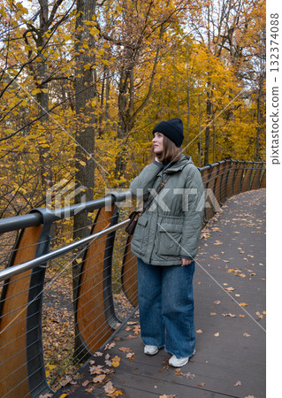 Woman in a green quilted jacket and black knit hat walking on the wooden eco trail bridge at VDNH park, surrounded by golden autumn trees 132374088