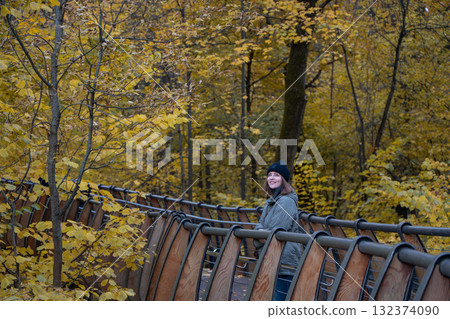Woman in a black beanie and green quilted jacket standing on the eco trail bridge at VDNH park, surrounded by golden autumn trees Woman in a black beanie and green quilted jacket standing on the eco trail bridge at VDNH park, surrounded by golden autumn trees 132374090