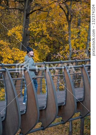 Woman in a green quilted jacket and black beanie smiles while standing on the eco-trail bridge at VDNH, surrounded by yellow autumn trees Woman in a green quilted jacket and black beanie smiles while standing on the eco-trail bridge at VDNH, surrounded by yellow autumn trees 132374104