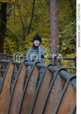 Woman in a black beanie and green quilted jacket standing on the eco trail bridge at VDNH park, surrounded by golden autumn trees 132374106