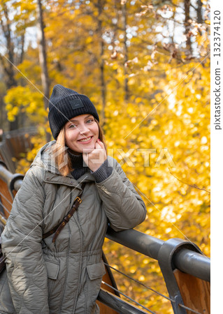 Smiling woman in a green quilted jacket and black knit hat leaning on a wooden bridge surrounded by golden autumn trees Smiling woman in a green quilted jacket and black knit hat leaning on a wooden bridge surrounded by golden autumn trees 132374120