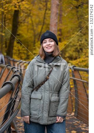 Woman in a green quilted jacket and black knit hat smiling on the eco trail bridge at VDNH park, surrounded by autumn trees Woman in a green quilted jacket and black knit hat smiling on the eco trail bridge at VDNH park, surrounded by autumn trees 132374142