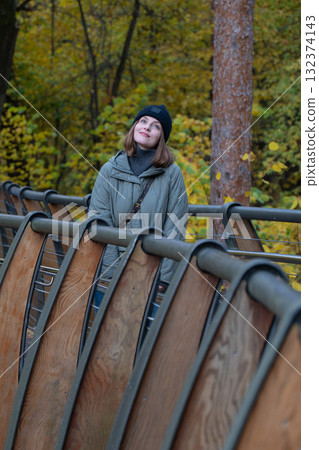 Woman in a black beanie and green quilted jacket standing on the eco trail bridge at VDNH park, surrounded by golden autumn trees 132374143