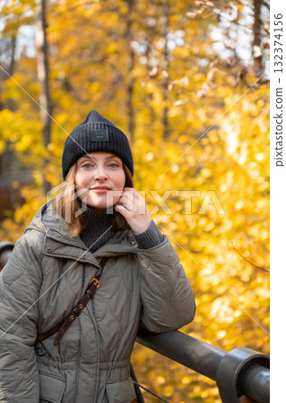 Woman in a green quilted jacket and black knit hat leaning on a wooden bridge surrounded by bright yellow autumn trees Woman in a green quilted jacket and black knit hat leaning on a wooden bridge surrounded by bright yellow autumn trees 132374156