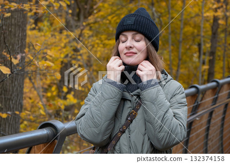 Woman in a black knit hat and green quilted jacket enjoying a peaceful autumn moment on the eco trail bridge at VDNH park Woman in a black knit hat and green quilted jacket enjoying a peaceful autumn moment on the eco trail bridge at VDNH park 132374158