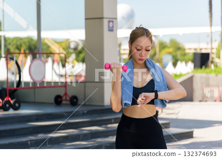 Woman sport exercise workout outdoor, fit people stretch. Young woman checks smartwatch during training, holding dumbbell for strength, enjoys active healthy lifestyle, prepares to stretch body again 132374693