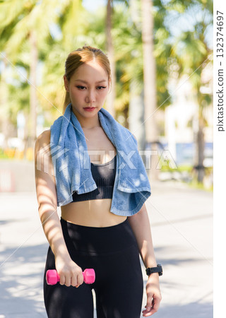 Stretch helps fit sport woman for outdoor workout, preparing for exercise Woman holds dumbbell, blue towel over shoulders, performing warm up stretch for fitness, enjoying sunny day 132374697