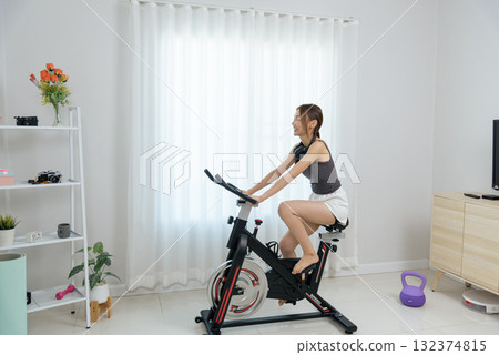Young happy woman active at home, people enjoy fitness workout on indoor cycling bike wearing headphones, healthy lifestyle activity promoting wellness in bright home environment Young happy woman active at home, people enjoy fitness workout on indoor cycling bike wearing headphones, healthy lifestyle activity promoting wellness in bright home environment 132374815