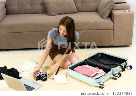 Young woman at home, happy people enjoy activity, meticulously packing travel luggage with clothes camera laptop planning exciting vacation journey 132374825