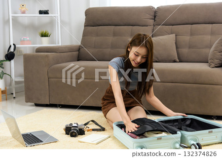 Young happy woman at home, displaying common people activity for travel planning, packing clothes into light blue suitcase, preparing for vacation adventure with laptop and camera ready 132374826