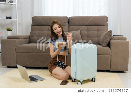 Young happy woman at home plans travel activity, writing trip plans in notebook, enthusiastically preparing for exciting vacation with camera laptop phone and light blue suitcase 132374827