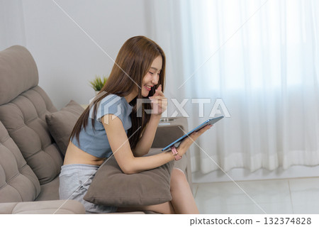 Young happy woman at home enjoying casual activity, people find joy with modern technology, she relaxes comfortably on couch, engaging with digital tablet, feeling cheerful and connected 132374828