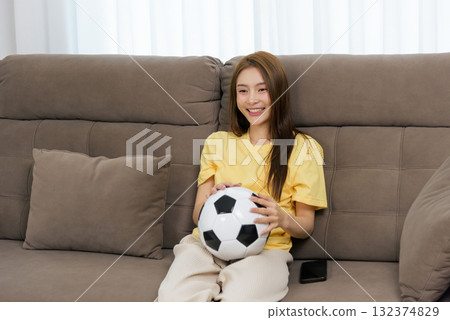 Young happy woman enjoys home activity, people like her watch football, holding soccer ball on comfortable sofa for relaxation indoors 132374829