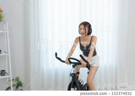 Young woman at home enjoys happy exercise activity, people see her smiling brightly, cycling indoor on stationary bike for fitness, listening to music with headphones, promoting healthy lifestyle 132374832