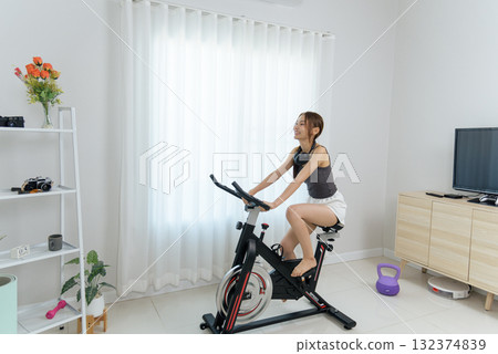 Young happy woman pedals spin bike for home fitness activity, people enjoying healthy exercise inside bright room displaying white curtains and shelf with kettlebell 132374839