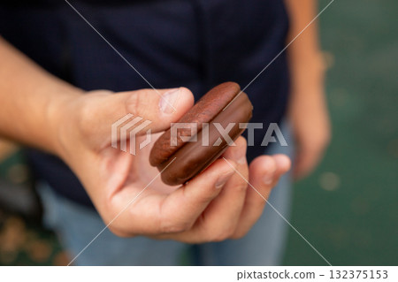 Two layers of sponge cake with a marshmallow or souffle layer, covered in chocolate glaze. round sponge cake with chocolate or chocolate cookie in man hand, close up view. 132375153