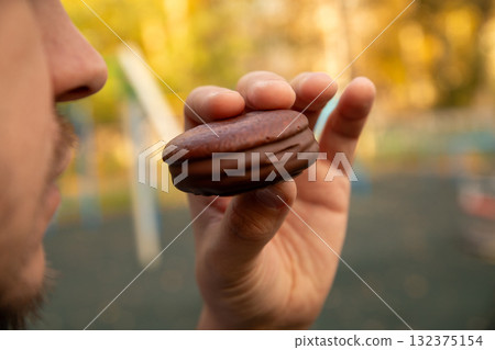 Airy sponge cake with delicate cream inside, covered in chocolate glaze. round chocolate cookie in man hand. Airy sponge cake with delicate cream inside, covered in chocolate glaze. round chocolate cookie in man hand. 132375154