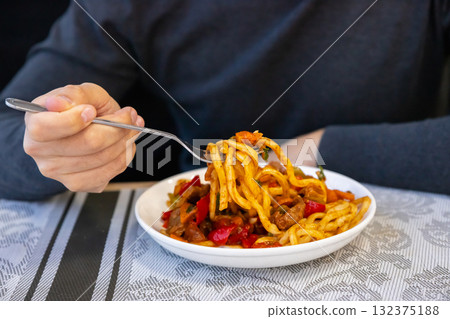 Lagman noodles. A hand holds a fork with a generous portion of pasta, topped with colorful vegetables. The cozy restaurant setting adds to the warm lunchtime atmosphere where meals are enjoyed. Lagman noodles. A hand holds a fork with a generous portion of pasta, topped with colorful vegetables. The cozy restaurant setting adds to the warm lunchtime atmosphere where meals are enjoyed. 132375188