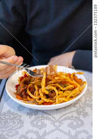 hand holds fork with generous portion of pasta, topped with colorful vegetables. cozy restaurant setting adds to warm lunchtime atmosphere where meals are enjoyed. Lagman noodles. hand holds fork with generous portion of pasta, topped with colorful vegetables. cozy restaurant setting adds to warm lunchtime atmosphere where meals are enjoyed. Lagman noodles. 132375189