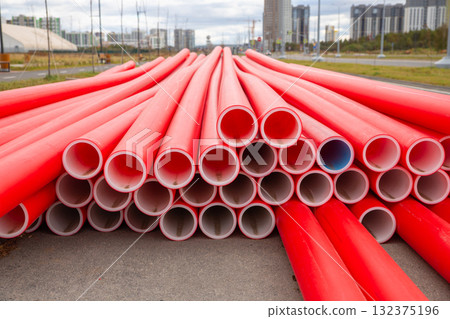 Stack of bright red plastic conduit pipes is arranged in a neat stack on a construction site. Modern buildings rise in the background under a partly cloudy sky, indicating urban development. 132375196