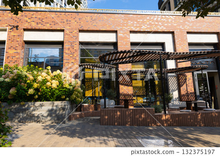 A cozy outdoor seating area of cafe or restaurant features stylish furniture under a canopy in a vibrant urban environment. Lush plants and flowers enhance the inviting atmosphere on a sunny day. A cozy outdoor seating area of cafe or restaurant features stylish furniture under a canopy in a vibrant urban environment. Lush plants and flowers enhance the inviting atmosphere on a sunny day. 132375197