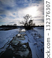 Cracked ice and a river bed, stream with a tree silhouette at sunset 132376307