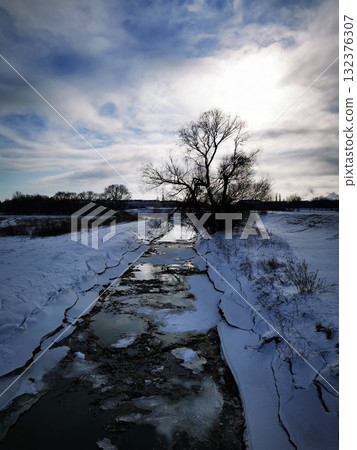 Cracked ice and a river bed, stream with a tree silhouette at sunset 132376307