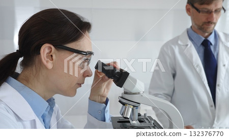 A female scientist wearing protective glasses looks into a microscope and conducts a medical research in a sterile laboratory while her male colleague works in the background. Medicine and science 132376770