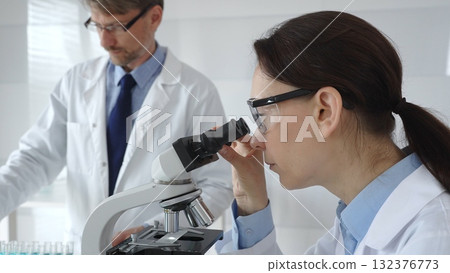 Female scientist wearing protective glasses looking through a microscope during medical research in laboratory with her male colleague working in the background. Medicine and science concept Female scientist wearing protective glasses looking through a microscope during medical research in laboratory with her male colleague working in the background. Medicine and science concept 132376773