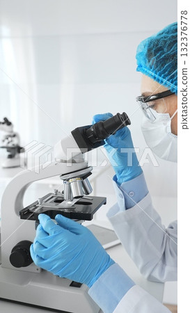 Female scientist wearing blue protective gear using a microscope examining samples in a laboratory setting, conducting scientific research and analysis. Medicine and science concept Female scientist wearing blue protective gear using a microscope examining samples in a laboratory setting, conducting scientific research and analysis. Medicine and science concept 132376778