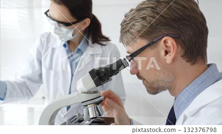 A researcher wearing glasses examines a sample under a microscope, a lab technician wearing a protective mask stands nearby, both in white lab coats during the study. Medicine and Science A researcher wearing glasses examines a sample under a microscope, a lab technician wearing a protective mask stands nearby, both in white lab coats during the study. Medicine and Science 132376798