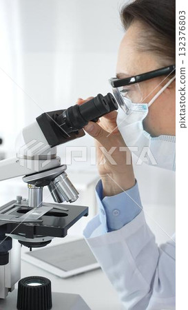 A female scientist in a protective mask and glasses carefully examines a sample under a microscope while conducting research in a modern laboratory. Concept of medicine and science A female scientist in a protective mask and glasses carefully examines a sample under a microscope while conducting research in a modern laboratory. Concept of medicine and science 132376803