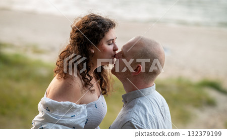Couple sharing a romantic kiss on a beach during sunset near calm waters and soft grass Couple sharing a romantic kiss on a beach during sunset near calm waters and soft grass 132379199