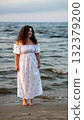 Woman in a white off-shoulder dress standing by the water at sunset on a beach 132379200