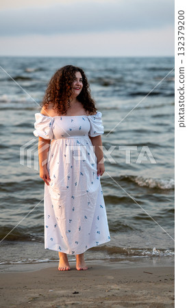 Woman in a white off-shoulder dress standing by the water at sunset on a beach Woman in a white off-shoulder dress standing by the water at sunset on a beach 132379200