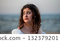 Young woman with curly hair gazes thoughtfully at the ocean during a serene beach sunset 132379201