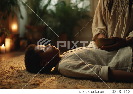 A child lies on a soft surface, eyes closed, as an adult gently places their hands on the child's head, surrounded by lit candles and plants, creating a peaceful atmosphere 132381140