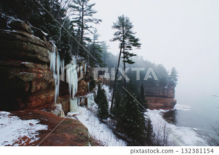 Icicles of various sizes dangle from the rocky cliffs in a forest during winter. The ground is covered in ice and there is a serene blue hue among the frozen landscape 132381154