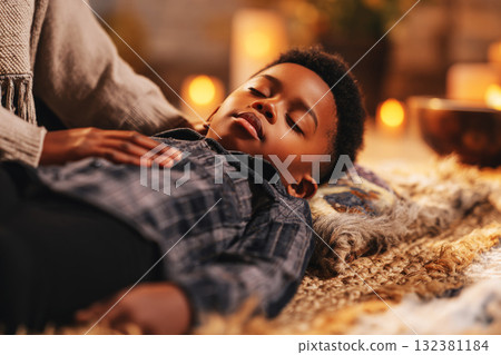 A child lies on a soft surface, eyes closed, as an adult gently places their hands on the child's head, surrounded by lit candles and plants, creating a peaceful atmosphere 132381184