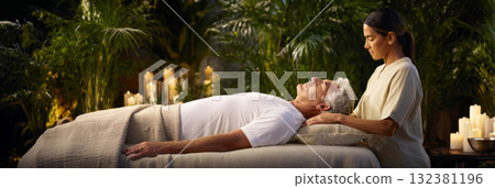 A person receives sound therapy while lying on a massage table surrounded by lush greenery and glowing candles, creating an atmosphere of relaxation and peace during the evening hours, banner 132381196