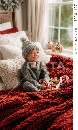 A happy toddler in a festive Santa hat sits on a soft red blanket, laughing with joy. Candy canes are nearby, creating a warm and cheerful holiday atmosphere 132381197