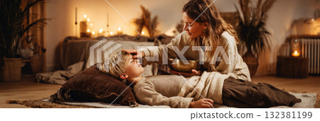 Child lies peacefully on a soft pillow during a sound therapy session, as a practitioner holds a singing bowl, creating soothing sounds in a serene space filled with greenery, banner 132381199