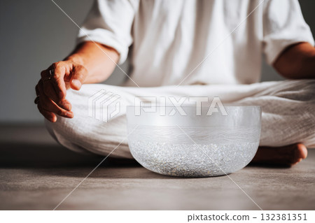 Practitioner sits calmly in a peaceful environment, focusing on sound therapy through a singing bowl while meditating. Soft, swirling waves of sound surround him, enhancing relaxation 132381351
