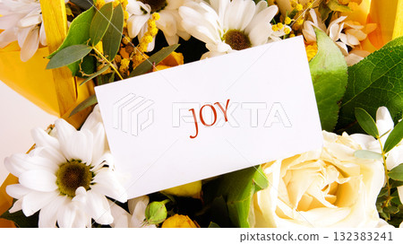A vibrant bouquet filled with white daisies and yellow roses features a card displaying the word joy. This beautiful arrangement radiates happiness and warmth 132383241