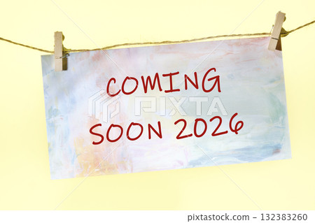 A vibrant sign displays the words coming soon 2026, hanging lightly from two wooden clips on a string, set against a pale yellow backdrop, creating an atmosphere of anticipation 132383260