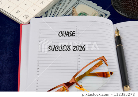 A detailed planner lays open on a blue surface showing the words chase success and the year 2026. Nearby, there are bills and a stylish pen, signaling financial planning and goal setting 132383278