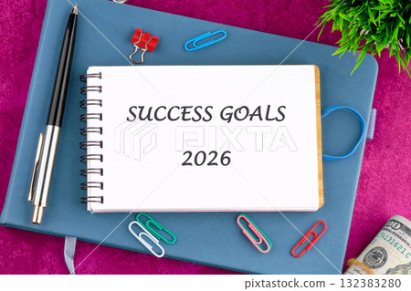 Notepad open to success goals for 2026 rests on a bright pink surface. Colorful paper clips, a pen, and a small plant create an inspiring workspace for future planning 132383280