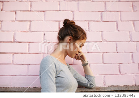 Young woman in fashion gray sweater with bun hair style standing near pink brick wall, expressing calmness, reflection and inner peace through minimalist lifestyle and balance. Casual street style Young woman in fashion gray sweater with bun hair style standing near pink brick wall, expressing calmness, reflection and inner peace through minimalist lifestyle and balance. Casual street style 132383442
