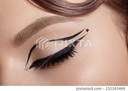 Close-up macro of a closed female eye with black winged eyeliner and tiny gold dot embellishment, soft lighting highlighting lash curve and eyelid decoration for editorial beauty use Close-up macro of a closed female eye with black winged eyeliner and tiny gold dot embellishment, soft lighting highlighting lash curve and eyelid decoration for editorial beauty use 132383469