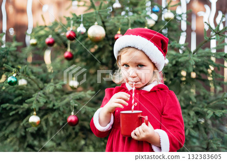 Little girl in red Santa dress and hat drinking from a red cup with a striped straw in front of a decorated outdoor Christmas tree 132383605